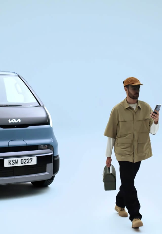 Dreiviertel-Frontansicht des Kia PV5 Passenger mit einem Mann, der ein Telefon und eine Lunchbox trägt und vorbeigeht.