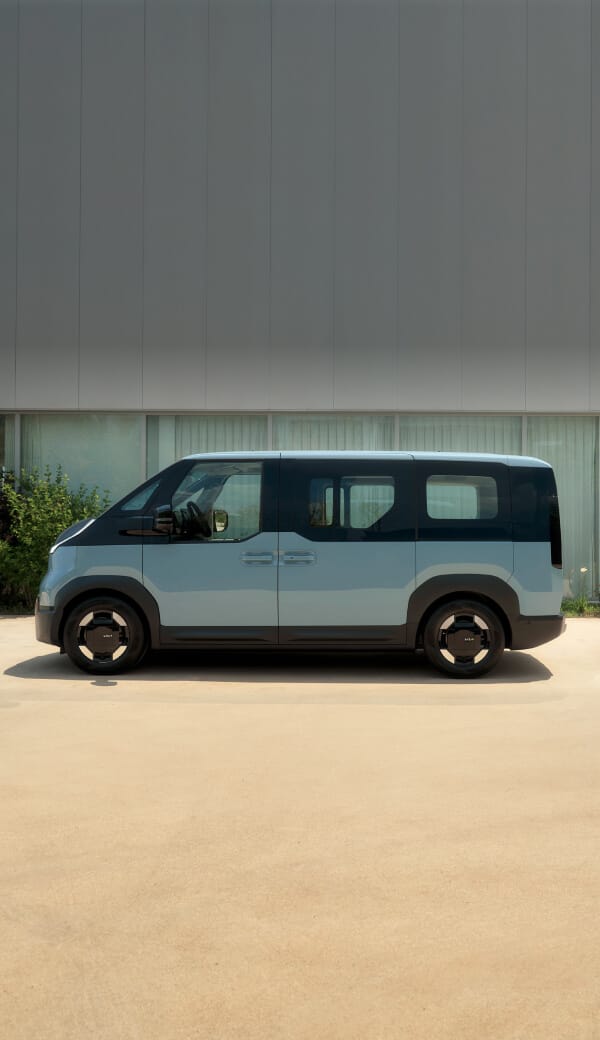 Vorderansicht des Kia PV5 Passenger, angeschlossen an eine Ladestation, mit einem Mann, der draußen neben Campingzubehör sitzt.